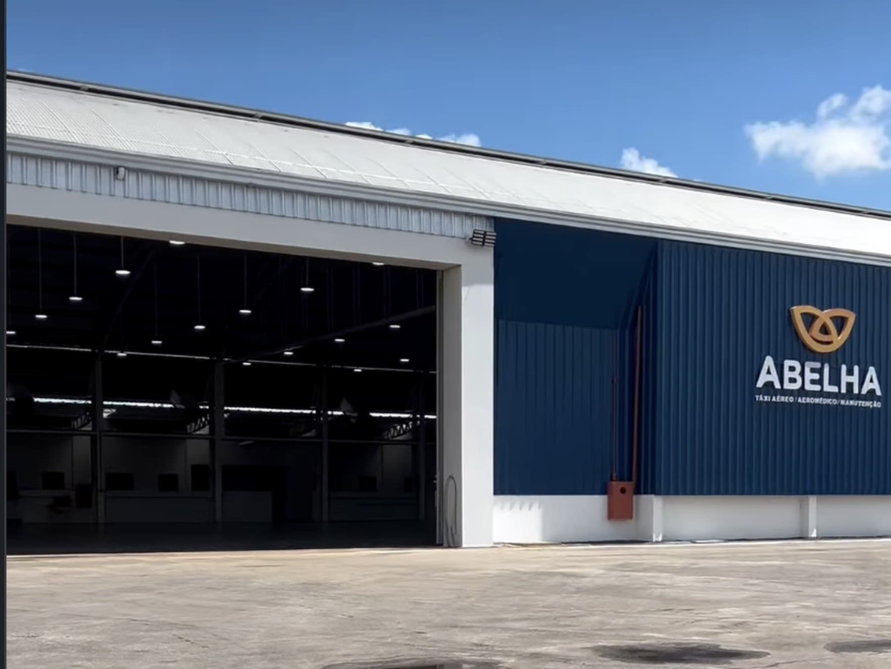 Hangar do Aeroporto Abelha, com o letreiro da empresa na fachada azul e o pátio em primeiro plano.